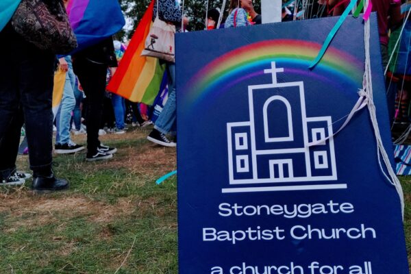Image shows a blue placard with the church logo under a rainbow and the words "Stoneygate Baptist Church: a church for all". The picture was taken at Leicester Pride 2022 and a number of people wearing pride flags can be seen in the background.