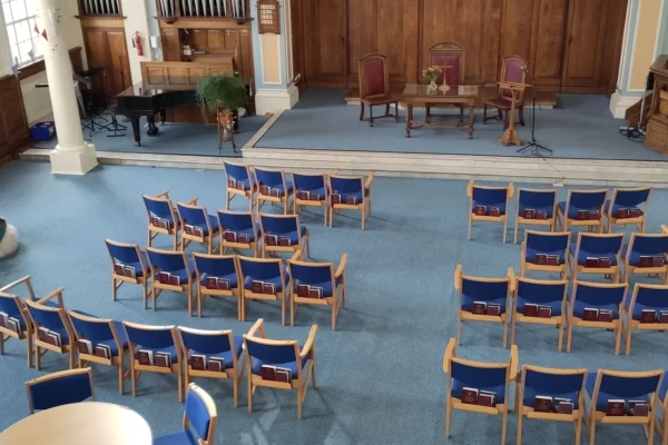 Image shows interior of main church space. The carpet is light blue and the walls are painted in gentle tones of peace and blue. Dark blue and wood chairs are laid out in rows and around tables. A raised platform at the front of the space holds a communion table and lectern. A piano and pipe organ are also visible.
