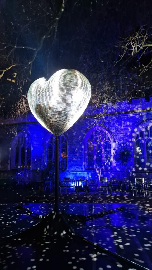 An uplit large disco ball heart, in front of the externals of a church building, lit up in blue.