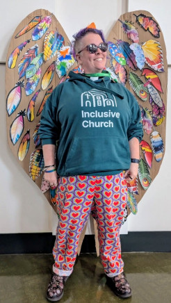 Grinning NatCo in a teal Inclusive Church hoodie and colourful trousers, infront of a set of colourful paper feathered wings. (The feathers are creative prayers from Generous Faith, coordinated by Emma Major)