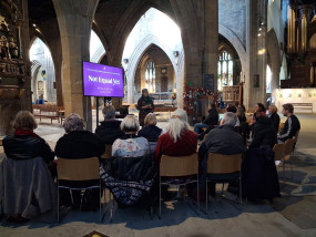 Group of people, mainly women, in a semi-circle facing a screen with 'Not Equal Yet' on it, there are arches and stained glass in the background, reflecting the event is in a church building.